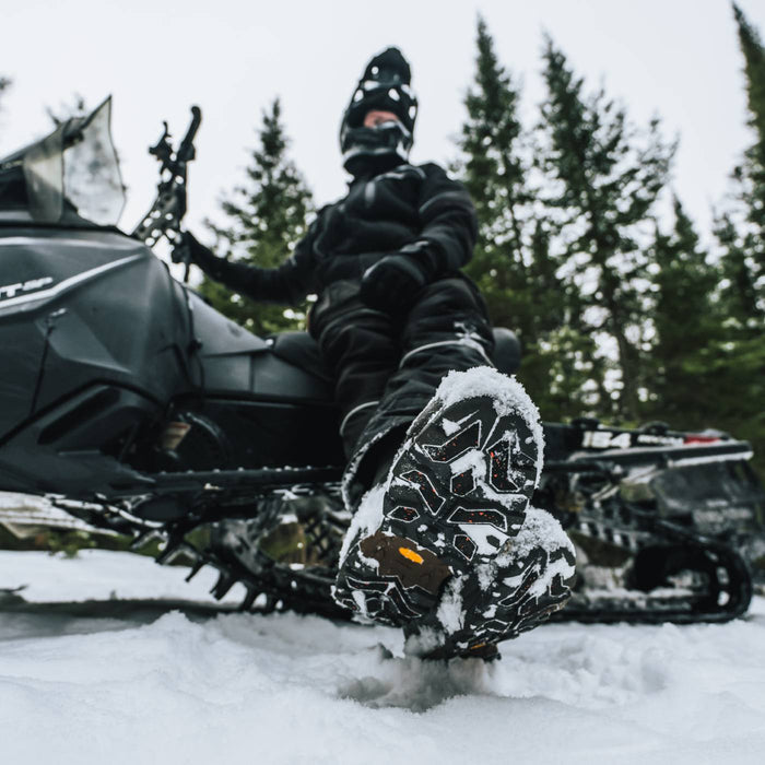 A person in black snowmobile riding gear sits on a black snowmobile in the snow, with one foot raised to show off snowboard boots covered in snow. Pine trees are visible in the background.