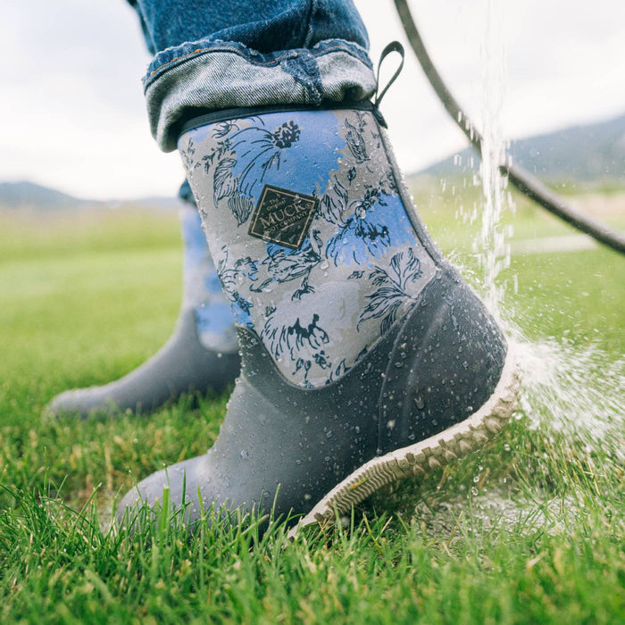 A close-up of a person wearing rubber rain boots with a blue and gray toile-style floral print on the upper and a dark gray rubber lower, standing on grass while water sprays from a hose.