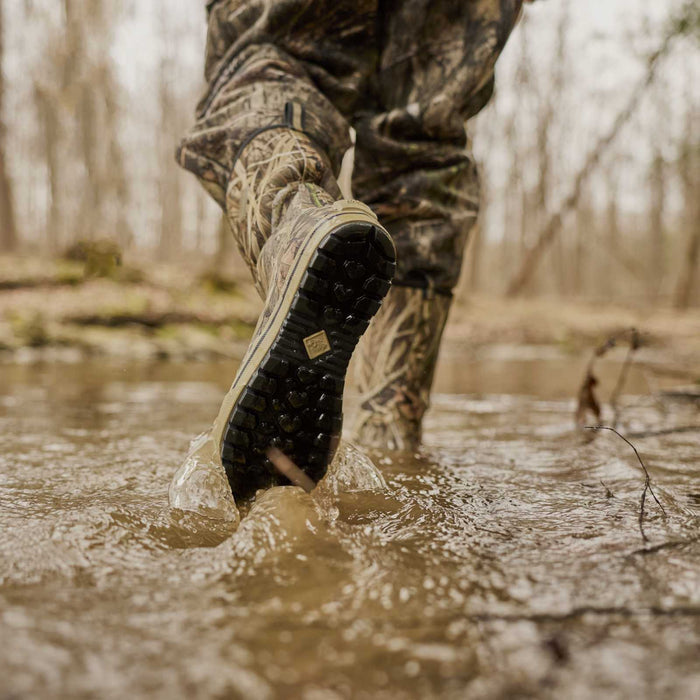 A close-up action shot of a person wading through shallow muddy water, showing the sole of a camouflage hunting boot with a black treaded outsole. The second boot is visible in the background.