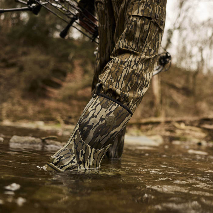 Person wearing Xplorer 12" Short Waterproof Rubber Boots in Mossy Oak Original Bottomland camo print walking through a shallow creek wearing hunting gear.