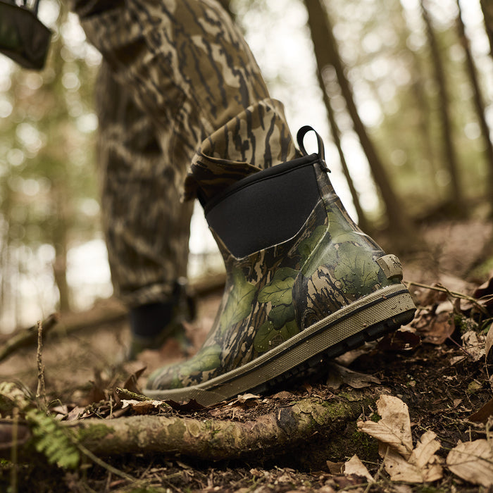 Camouflage boot on a forest floor with a person in camouflage clothing in the background.