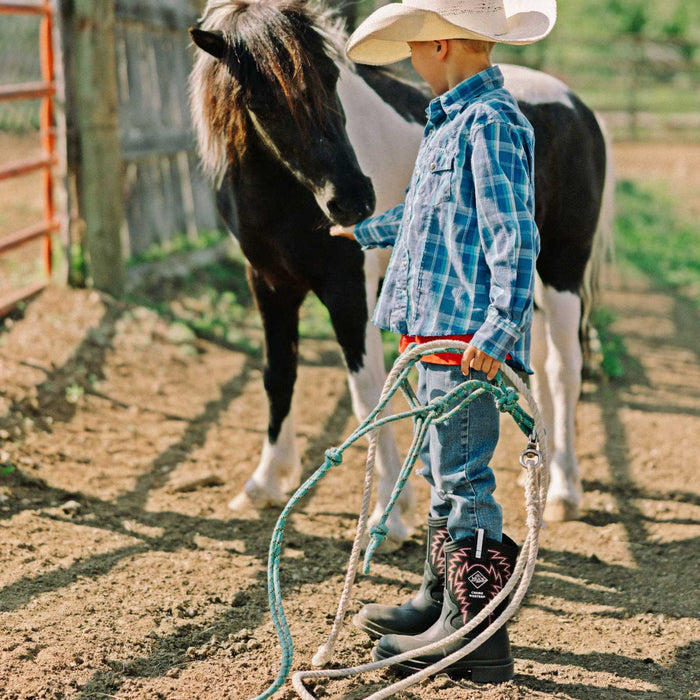Child in cowboy hat and boots standing next to a horse in a fenced area
