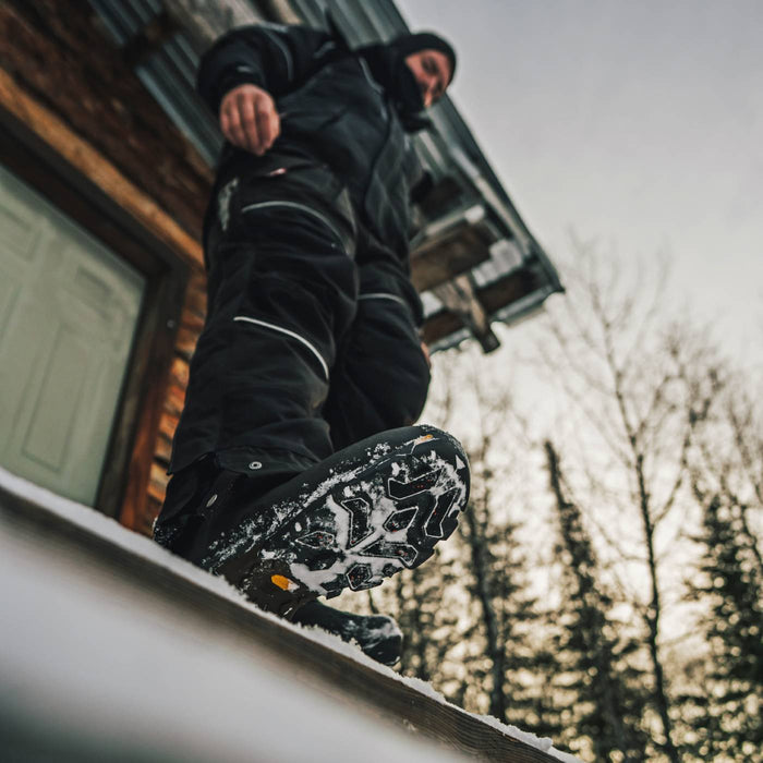 A person in black winter clothing steps onto a snowy roof in front of a wooden building, with bare trees and an overcast sky in the background. The product appears to be snowboard boots or winter footwear shown in use outdoors.