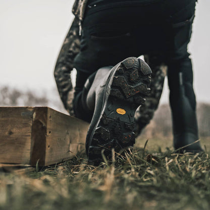 A close-up outdoor shot of a person wearing black rubber boots and dark pants, with one boot raised to show the tread and a small yellow Vibram logo on the outsole. The scene is set on grass beside a wooden plank.