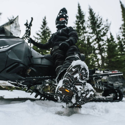 A person in black snowmobile riding gear sits on a black snowmobile in the snow, with one foot raised to show off snowboard boots covered in snow. Pine trees are visible in the background.