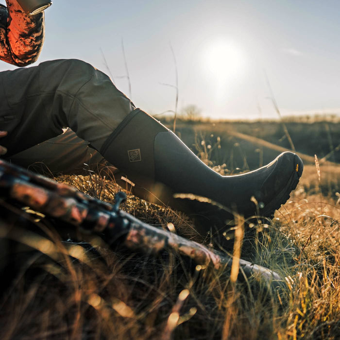 A person sitting in tall grass outdoors at sunset, wearing a dark rubber hunting boot and olive pants, with a camouflage-patterned rifle resting in the foreground.