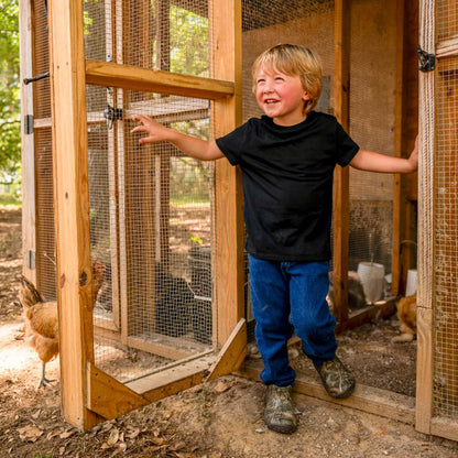 Child standing in a wooden chicken coop with chickens around wearing Mini Muck Kids Rain Boots in Mossy Oak Country DNA