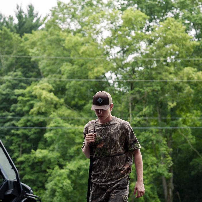 Muck® Diamond Trucker Hat, MACS900, Brown hat. Man in camouflage clothing and cap walking outdoors with trees in the background, Richardson Hats.