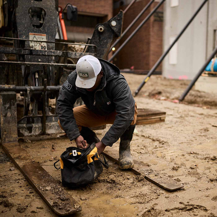 Muck® Logo Trucker Hat, Muck® Logo Trucker Hat, Gray hat, Man crouching getting tools out of tool bag. Richardson Hats.
