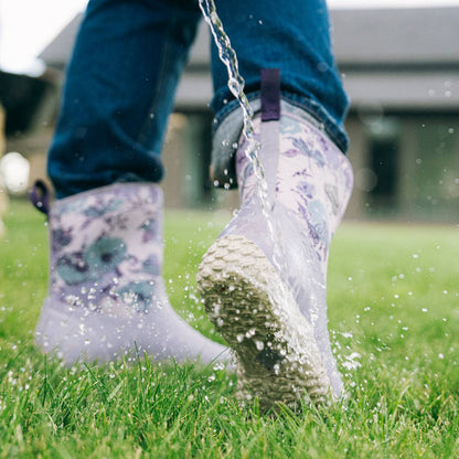 A person wearing floral-patterned rain boots walks on green grass with water splashing around them, paired with blue jeans.