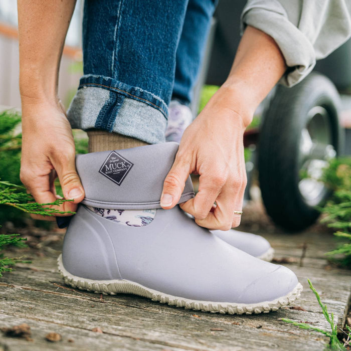 A person wearing a light gray Muck boot with blue jeans cuffed at the ankle, adjusting the boot on a rustic wooden surface outdoors.