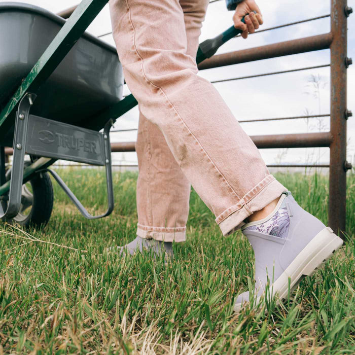 A person outdoors in a field wearing light gray slip-on boots with patterned socks and light pink jeans, standing near a wheelbarrow and metal fence.