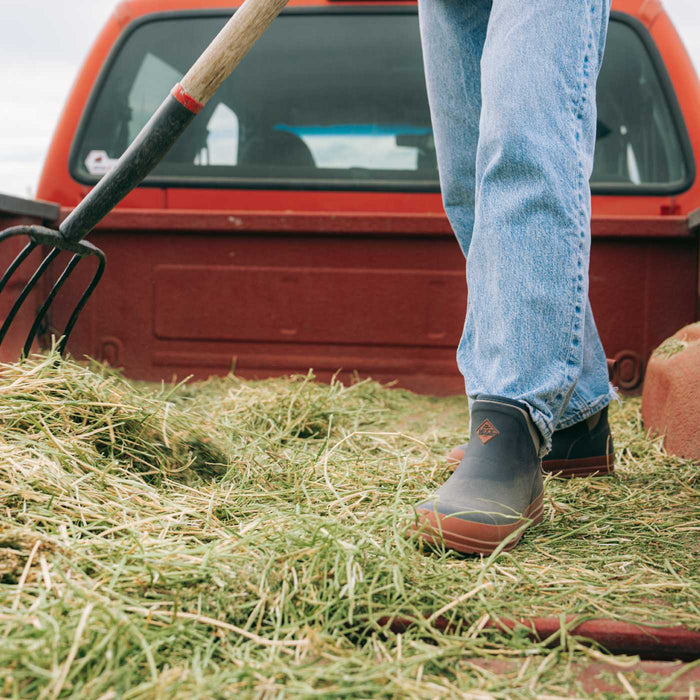 A person standing in a bed of hay next to a red pickup truck, wearing blue jeans and dark rubber boots, with a pitchfork visible in the foreground.