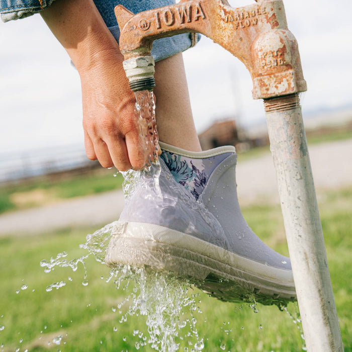 A person wearing Women's Originals Ankle in Minimal Gray Floral, rinsing it under running water from a rusted metal garden faucet outdoors on a grassy field.