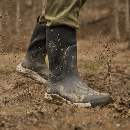 A close-up action shot of a person wearing dark gray rubber rain boots with mud splashing off them, standing in a muddy outdoor forest setting.