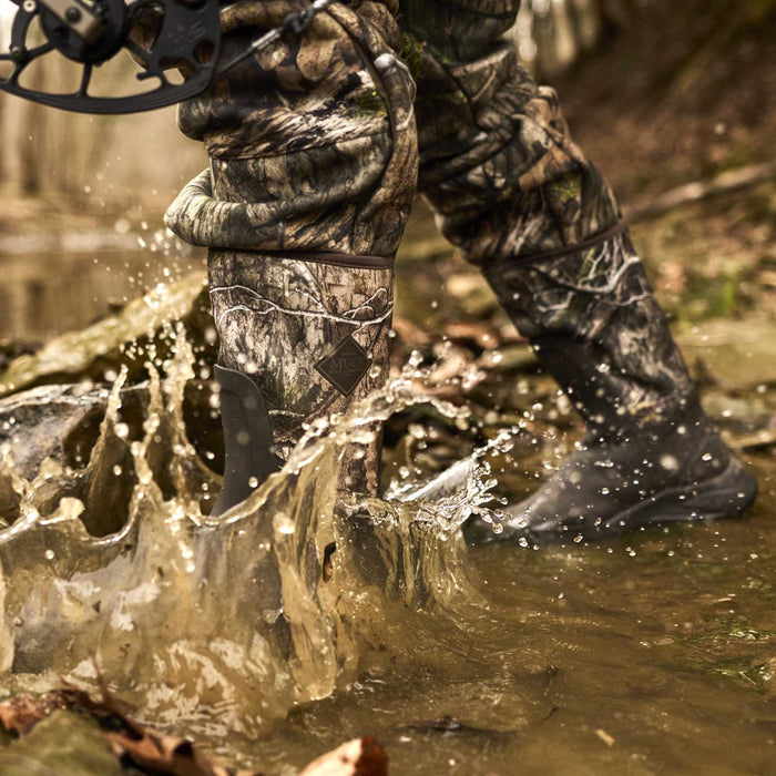 A close-up action shot of a person wearing camouflage hunting pants and black rubber boots, splashing through muddy water outdoors. A compound bow is partially visible in the upper left corner.