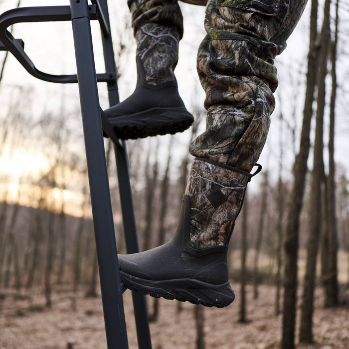 A person climbing a black metal ladder in a wooded outdoor setting, wearing camouflage hunting pants and black rubber boots.