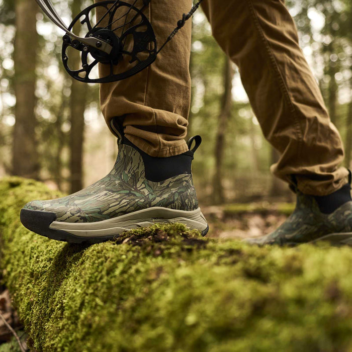 A close-up of a person wearing camouflage-patterned waterproof hiking boots with tan pants, standing on a moss-covered log in a forest while holding a bow compound bow.