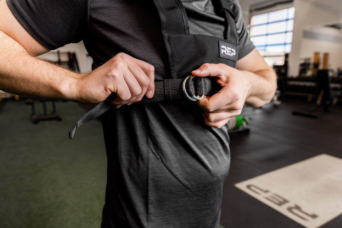 Person adjusting a black Sled Harness with a brand logo in a gym setting