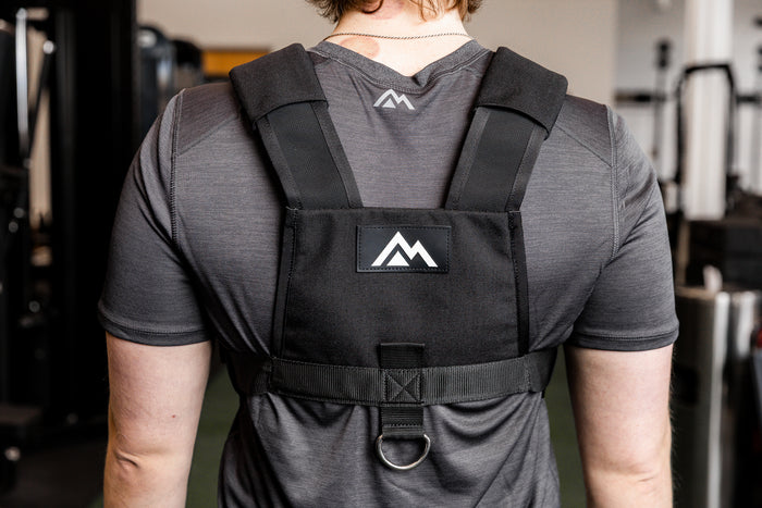 Person wearing a black Sled Harness with mountain logo in a gym setting