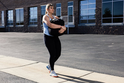 Woman performing a stone sandbag carry outdoors on a sidewalk with a building in the background
