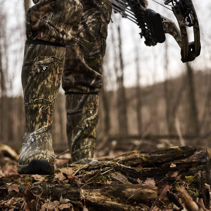 Close-up of a person wearing camouflage hunting boots and matching pants while standing on a log in a wooded outdoor setting, holding a compound bow.
