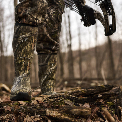 Close-up of a person wearing camouflage hunting boots and matching pants while standing on a log in a wooded outdoor setting, holding a compound bow.