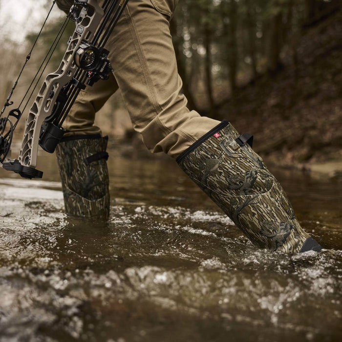 A person wading through shallow water in a forest while wearing tall camouflage hunting boots and tan pants, with a compound bow partially visible.