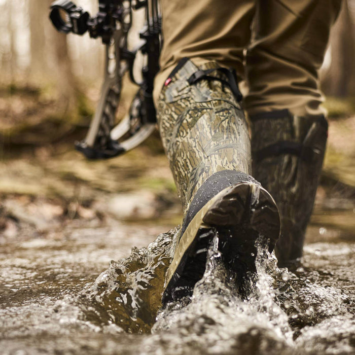 A close-up action shot of a person wading through shallow water in a forest, wearing camouflage hunting boots and tan pants, with a compound bow visible in the background.