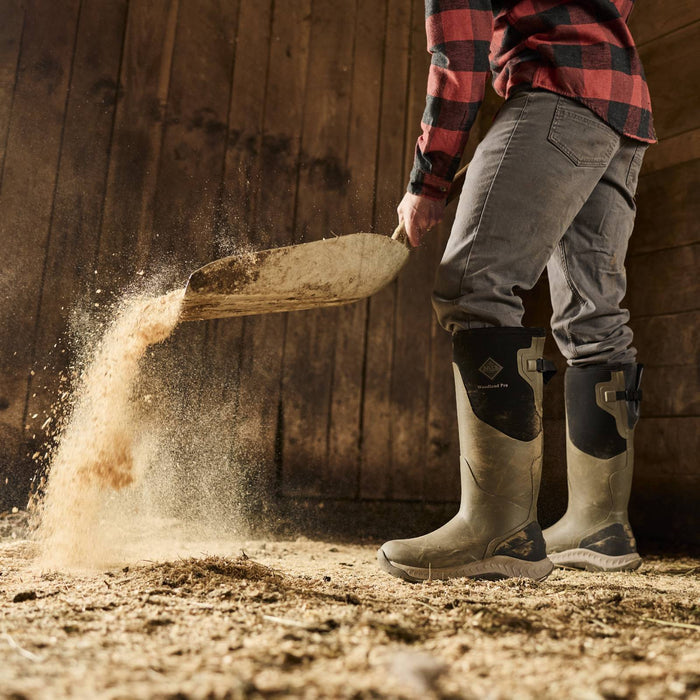 A person in a barn wearing tall gray and black boots while holding a shovel and pouring loose straw or manure onto the ground, with wood plank walls in the background.