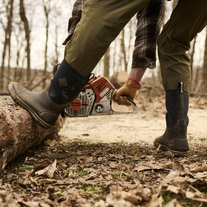 Person wearing Xplorer Men's 12" Short Rubber Boots while using a chainsaw to cut a fallen tree.