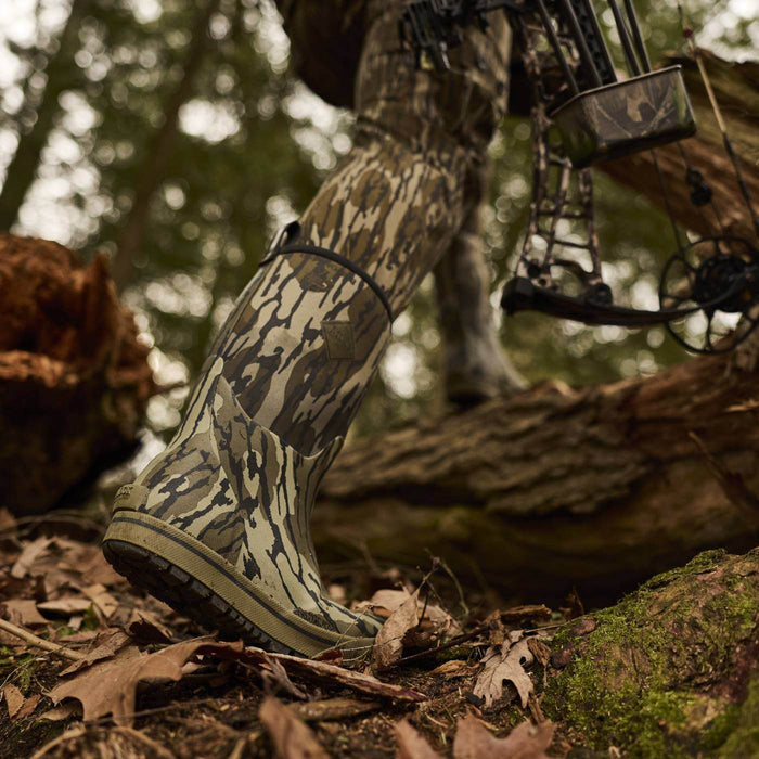 Person wearing camouflage hunting gear walking through woods with Xplorer 12" Short Waterproof Rubber Boots in Mossy Oak Original Bottomland camo print.