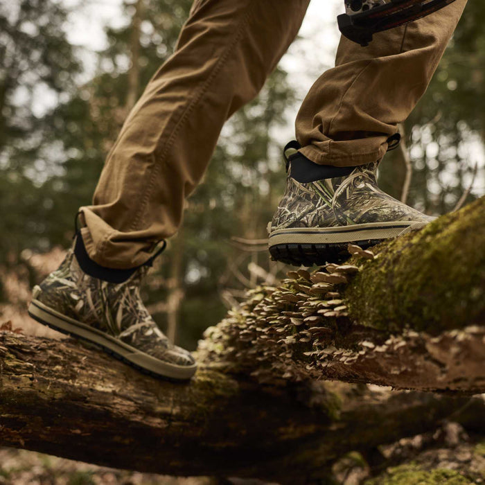 A close-up of a person's legs wearing camouflage hunting boots and tan pants while stepping carefully across a mossy log in a forest.