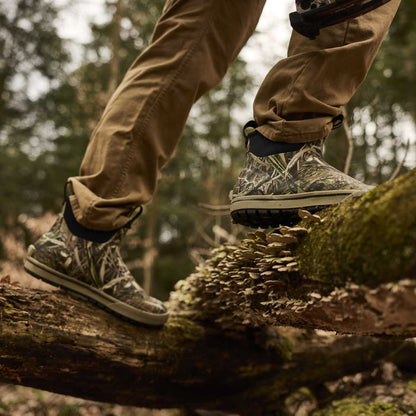 A close-up of a person's legs wearing camouflage hunting boots and tan pants while stepping carefully across a mossy log in a forest.
