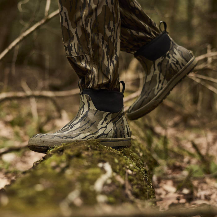 Person wearing camouflage pants and Mossy Oak Original Bottomland short  waterproof rubber camouflage boots walking on a mossy rock in a forest.