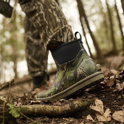 Camouflage boot on a forest floor with a person in camouflage clothing in the background.