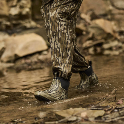 Person wearing camouflage pants and Xplorer Short Waterproof Rubber boots  in Mossy Oak Original Bottomland walking through water with rocks in the background